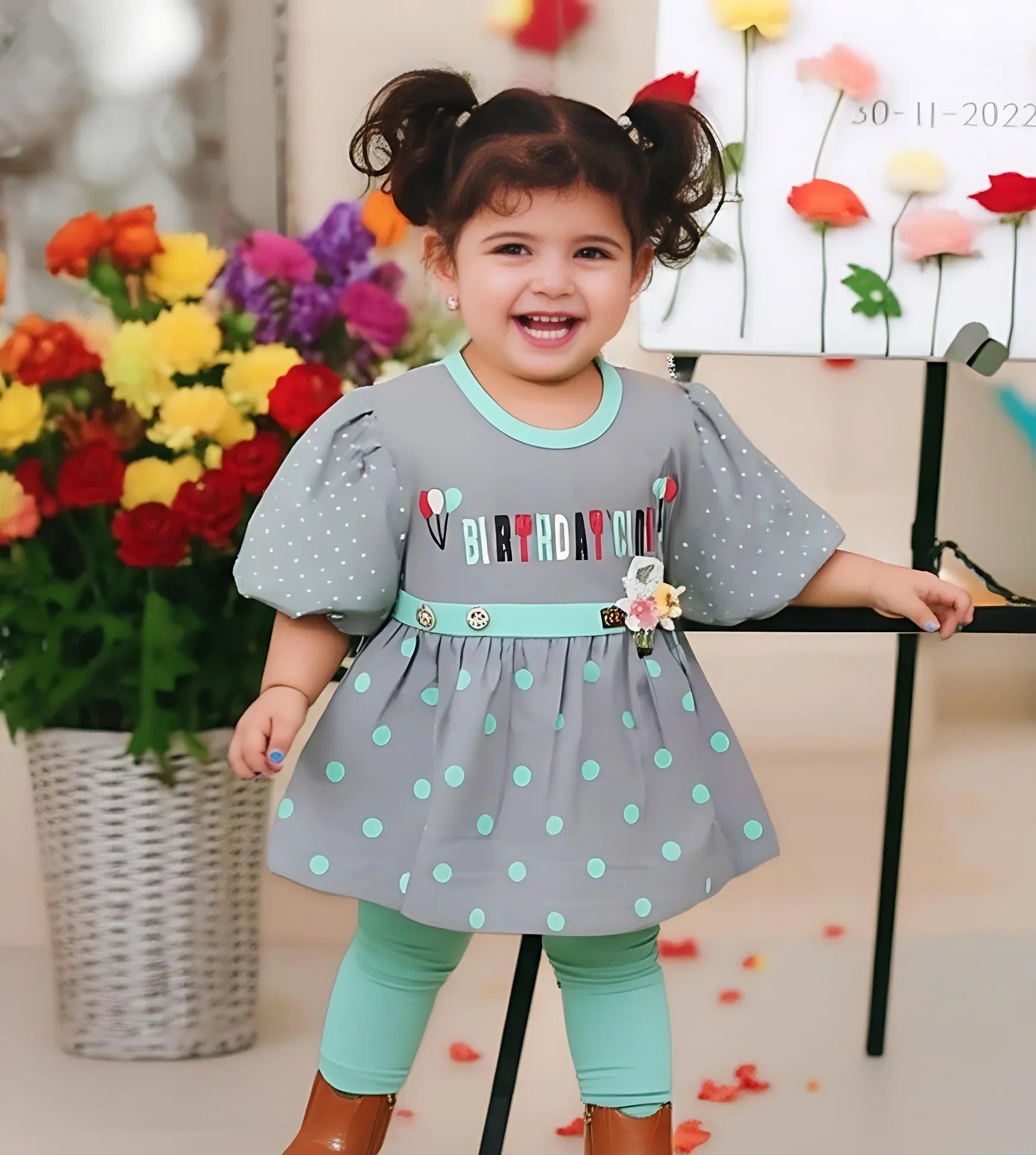 Child wearing a gray dress with polka dots and 'Happy Birthday' text, standing in front of flowers.