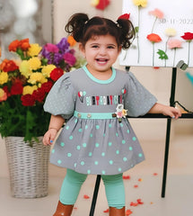 Child wearing a gray dress with polka dots and 'Happy Birthday' text, standing in front of flowers.