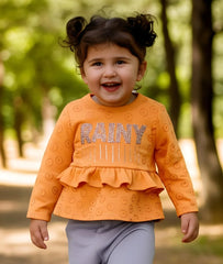Child wearing an orange shirt with 'RAINY' text, standing outdoors with trees in the background