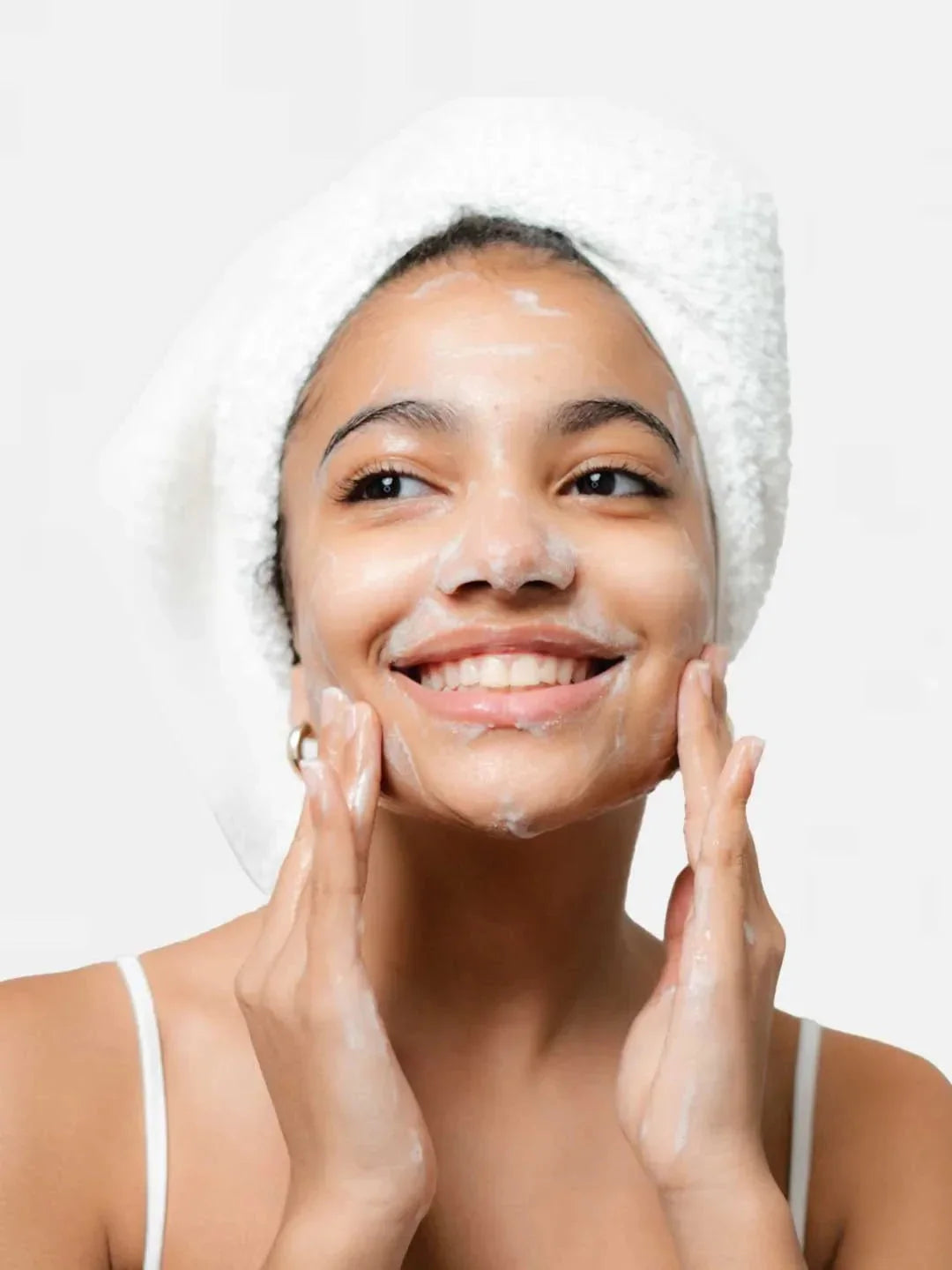 Woman applying cream to her face with a white towel on her head against a light background