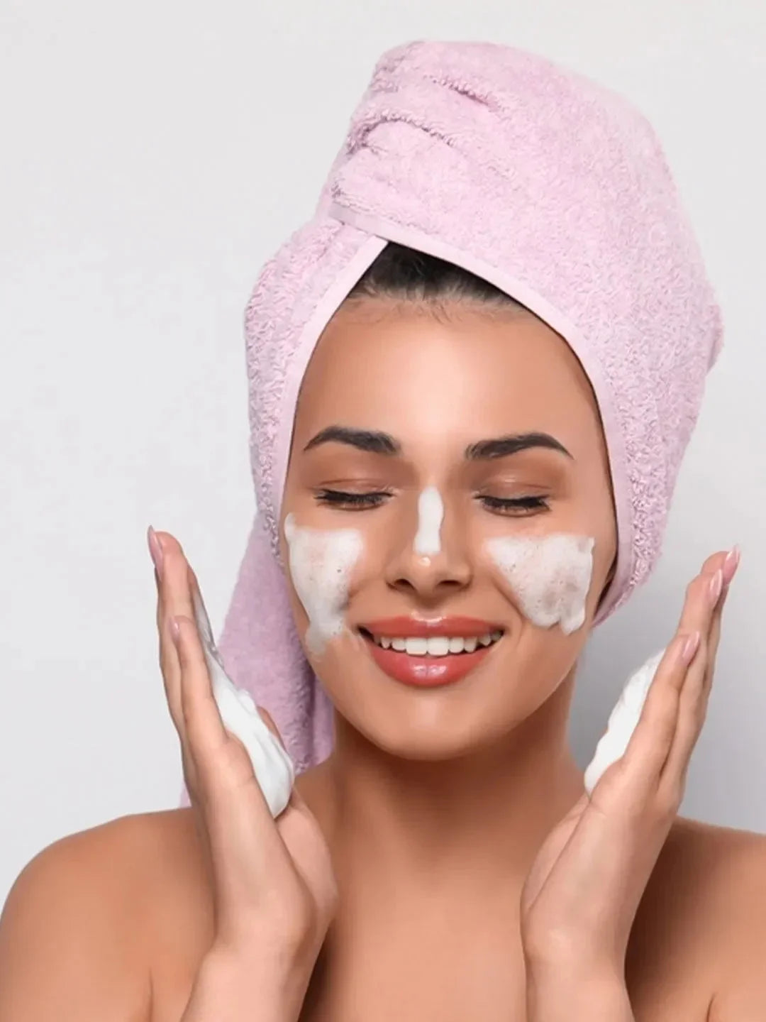 Woman applying facial mask with a pink towel on her head against a plain background