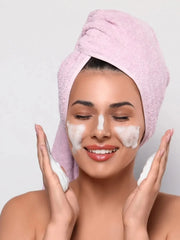 Woman applying facial mask with a pink towel on her head against a plain background